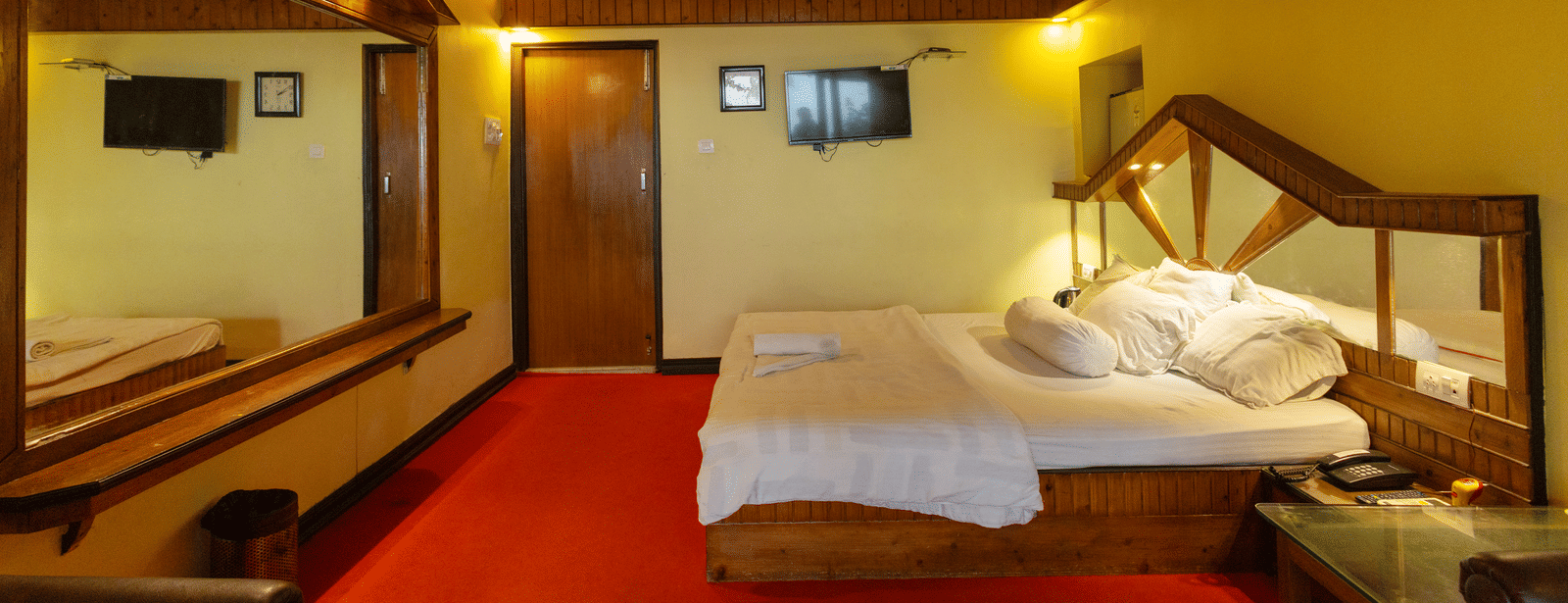 A bedroom with a king size bed, a wooden ceiling, a large mirror, a bedside table, and a wall mounted TV at Hotel Seagull Digha.